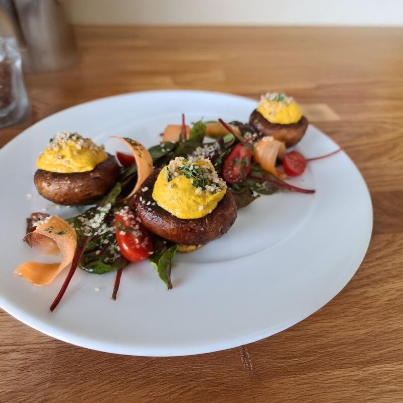 Gefüllte Champignons mit Muskat Haselnuss Karotte Drei gefüllte Champignons mit gelber Muskat-Haselnuss-Karotten-Creme stehen auf einem weißen Teller. Unterm Pilzhut liegen Blätter, Cherry-Tomaten und Karottenstreifen als Garnitur.