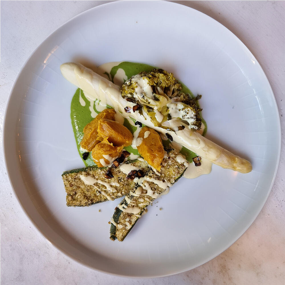 Spargel mit Hollandaise und Zucchini Schnitzel Weißer Spargel liegt auf grünem Püree, daneben sind zwei Scheiben Zucchini-Schnitzel mit heller Sauce. Auf dem Spargel und Schnitzeln ist cremige weiße Hollandaise verteilt.