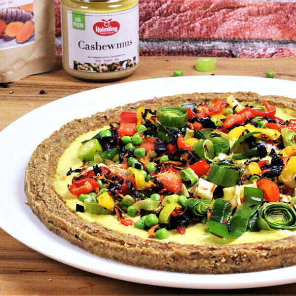 Indische Pizza mit Kurkumasoße, roh-vegan Eine Indische Pizza mit knusprigem Teig aus Mandeln, Flohsamenschalen- und Leinsamenmehl, belegt mit roten Zwiebeln, Lauch, Karotten, Paprika, Erbsen und einer cremigen gelben Kurkumasoße. Im Hintergrund steht ein Glas Cashewmus der Marke Keimling.