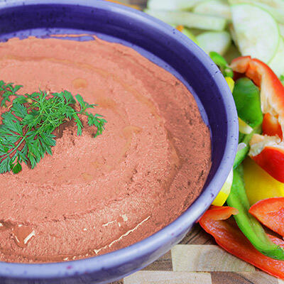 Paprika-Sesam-Aufstrich Cremiger Paprika-Sesam-Aufstrich in einer blauen Schale, garniert mit einem Zweig frischer Petersilie und umgeben von geschnittenen roten und grünen Paprikastreifen.