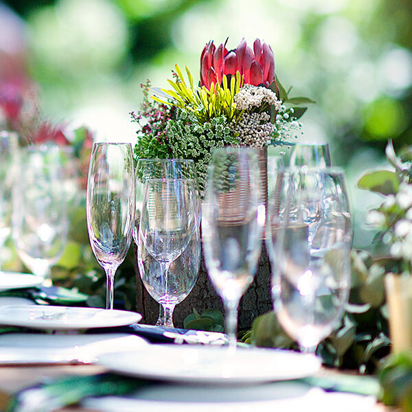 Vegane Sommergerichte von Boris Lauser Mehrere leere Weingläser und weiße Teller stehen auf einem gedeckten Tisch. Eine farbenfrohe Blumendekoration mit roten und weißen Blüten steht mittig auf dem Tisch. Der Hintergrund wirkt hell und unscharf.