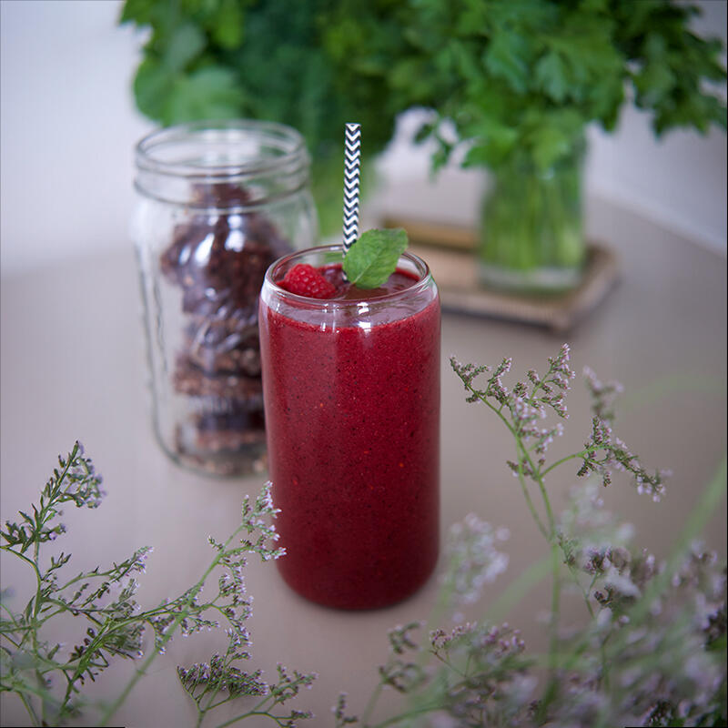 Beauty Smoothie von Maria Sperling Roter Smoothie mit Minzblatt und Himbeere im Glas mit Strohhalm steht auf dem Tisch. Im Hintergrund steht ein Glas mit Datteln. Vorn sind Pflanzenzweige mit kleinen lila Blüten.