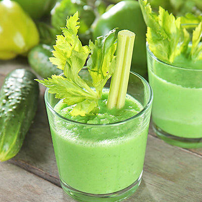 Stimmungsschmeichler Smoothie von Peter Dreverhoff Gläser mit grünem Smoothie, garniert mit Stielen und Blättern von Sellerie, stehen auf einem Holztisch. Im Hintergrund liegen frische Gurken und weitere grüne Zutaten.