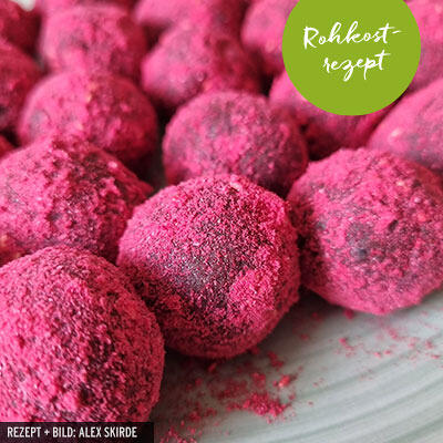 Runde Energiekugeln sind mit intensiv rotem Himbeerpulver bestäubt und liegen dicht nebeneinander auf einem weißen Teller. Ein grüner Sticker mit der Aufschrift Rohkostrezept ist oben rechts zu sehen.
