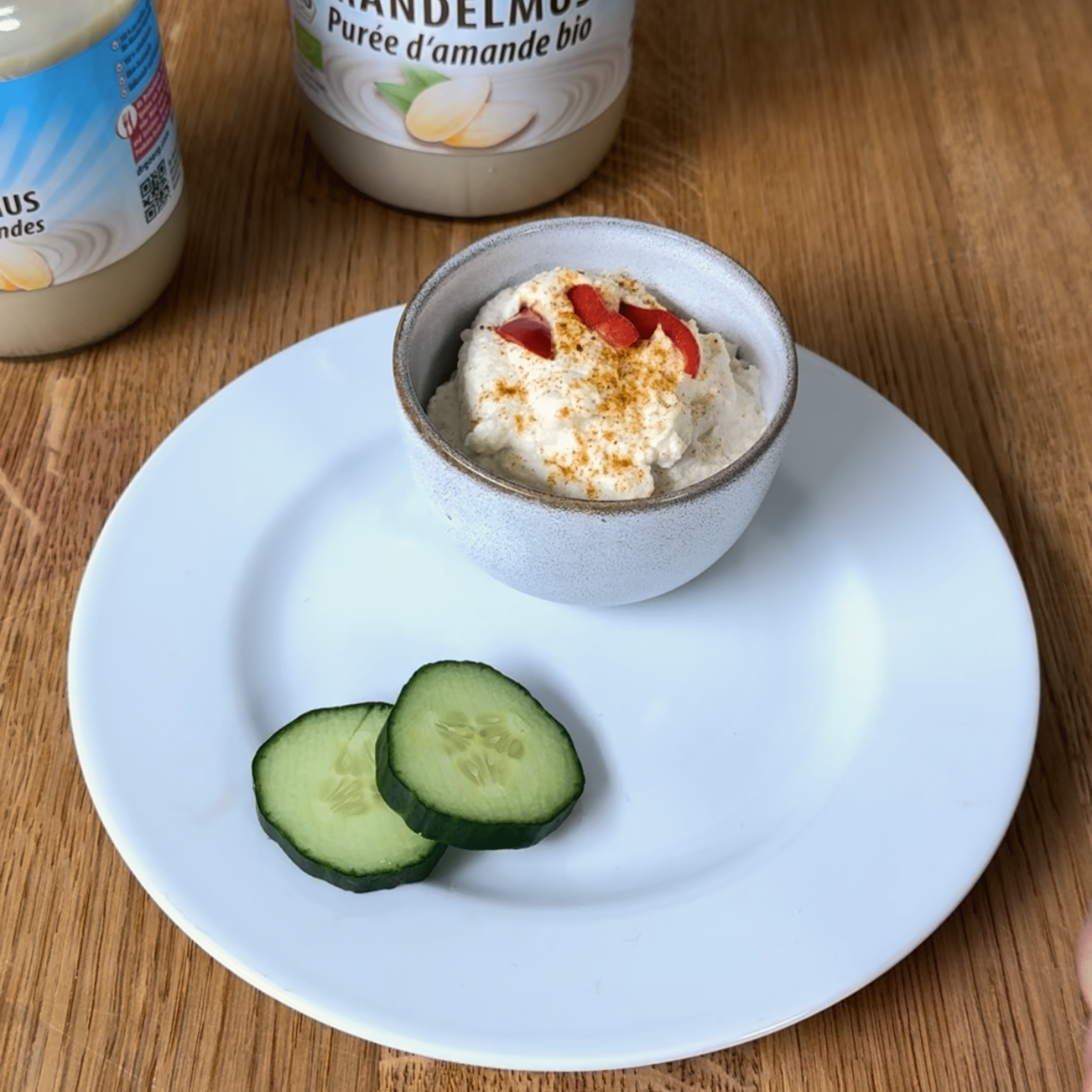 Feuriger Mandel Frischkäse Eine kleine Schale mit cremigem Frischkäse, bestreut mit roten Chilistücken und Gewürz, steht auf einem weißen Teller neben zwei Gurkenscheiben auf einem Holztisch.