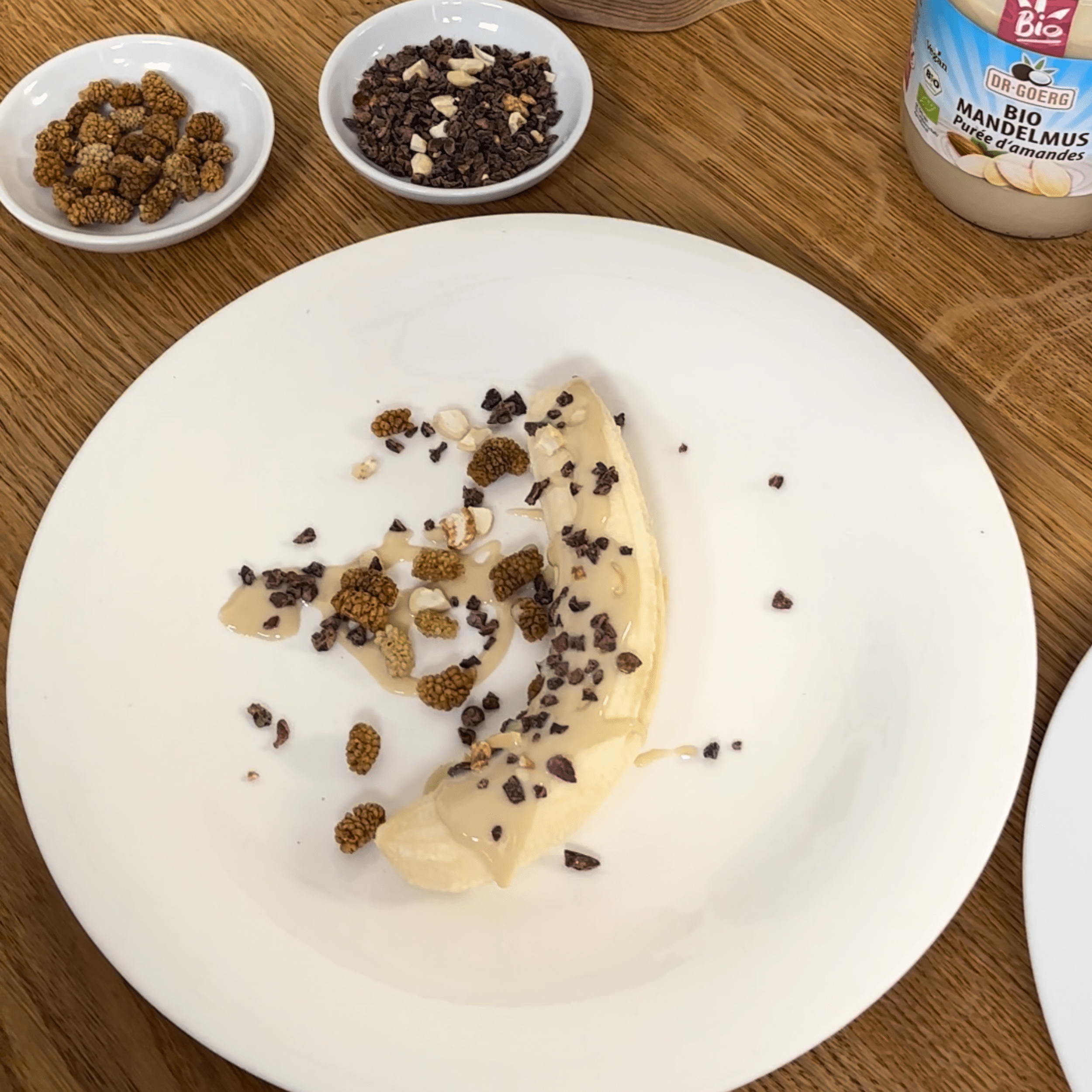 Schoko Bananen Flip  Halbierte Banane auf einem weißen Teller mit Mandelmus, Kakaonibs und getrockneten Maulbeeren als Topping. Daneben zwei kleine Schalen mit Kakaonibs und Maulbeeren sowie ein Glas Bio Mandelmus.