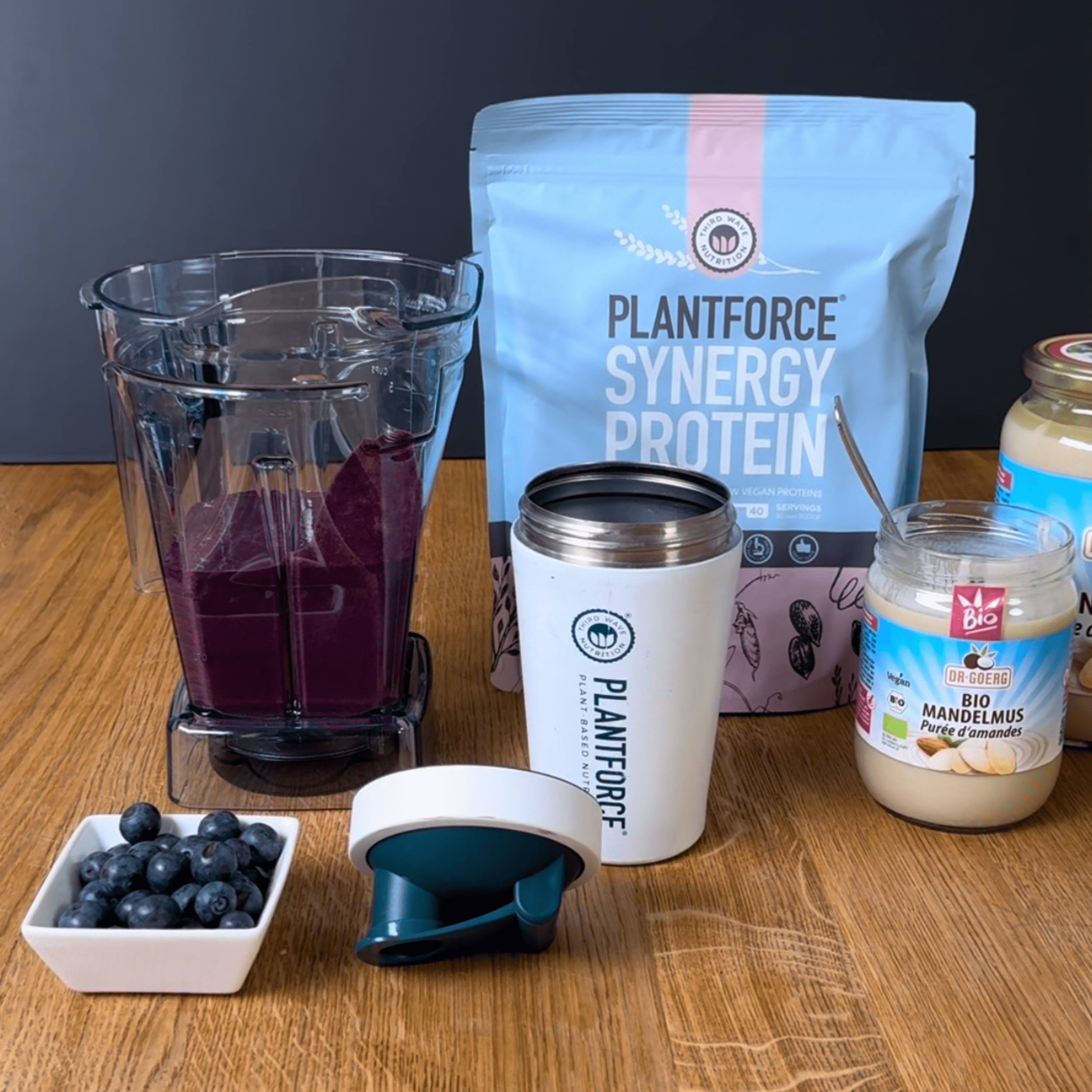 Mandel Proteinshake Packung Plantforce Synergy Protein steht neben einem offenen, weißen Proteinshake-Becher, einem Mixer mit lila Shake, einem Deckel und einem Glas mit Bio Mandelmus. Daneben liegt eine kleine Schale mit Heidelbeeren.