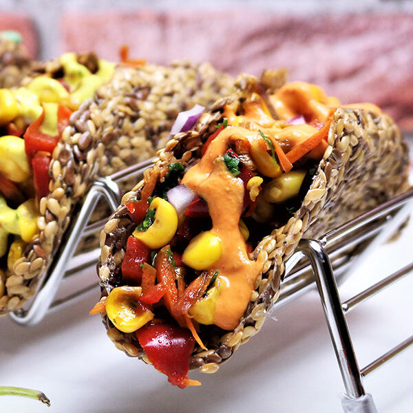 Party Tacos, roh-vegan Knusprige Tacos aus goldenen und braunen Leinsamen füllen sich mit Mais, roter Paprika, Karotten, Zwiebeln und grünen Kräutern. Cremige orange Soße läuft über das Gemüse.