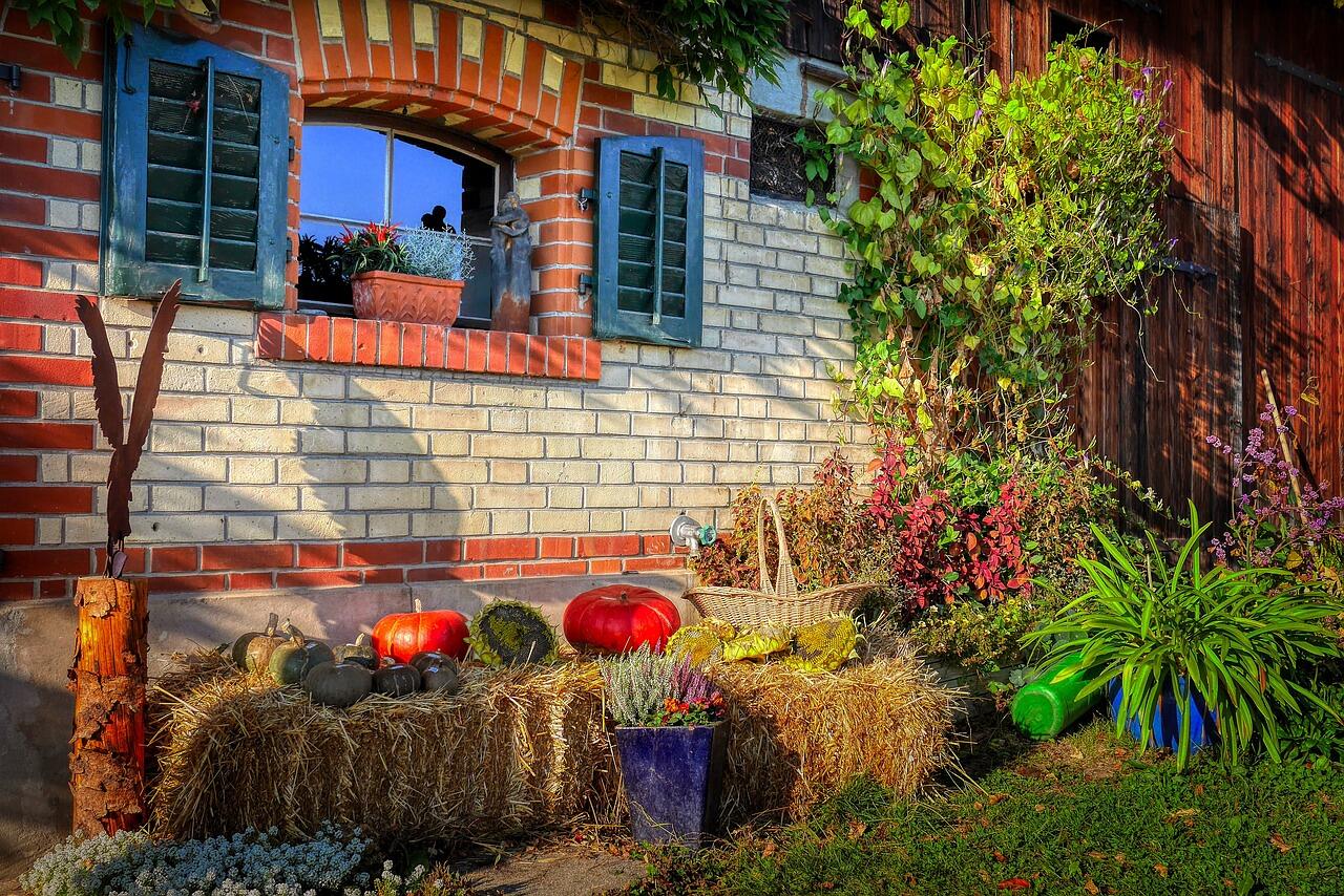 Zwei Heuballen mit verschiedenen Kürbissen, einem Korb, Blumentopf und Pflanzen stehen vor einer Hauswand mit geöffnetem Fenster und grünen Fensterläden.
