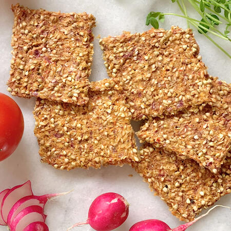 Fünf knusprige, quadratische Körnercracker liegen auf hellem Untergrund. Daneben sind frische Radieschen mit roter Schale und ein roter Tomate zu sehen.