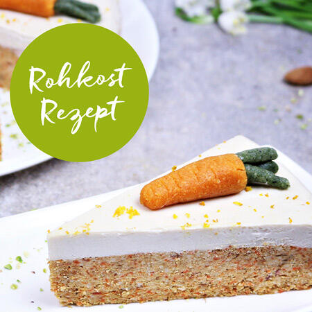 Ein Stück cremiger Rohkostkuchen mit weißer Frischkäse-Schicht und Marzipankarotte als Dekoration auf einem weißen Teller. Im Hintergrund steht ein weiterer Teller. Ein grüner Kreis zeigt den Text Rohkost Rezept.