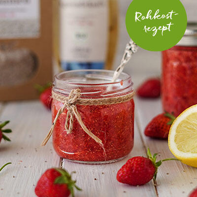 Erdbeer-Chia-Marmelade Glas mit roter Erdbeer-Chia-Marmelade, verziert mit einer Schnur, umgeben von frischen Erdbeeren und einer halben Zitrone auf hellem Holzuntergrund. Ein grüner Kreis zeigt den Schriftzug Rohkost-rezept.