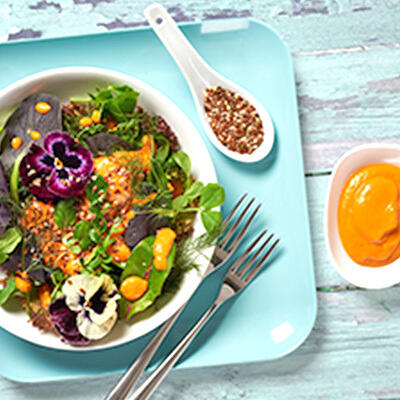 Paprika-Zucchini-Dressing Schüssel mit grünem Salat, essbaren Blüten und einem Paprika-Zucchini-Dressing, daneben Löffel mit Gewürzmischung und eine kleine Schale mit orangem Dressing auf hellblauem Tablett.
