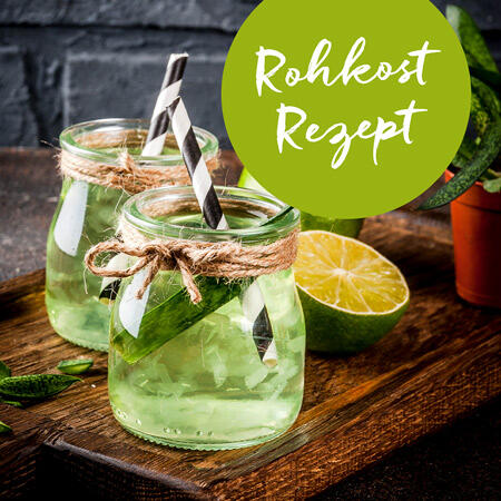Zwei Gläser mit grünem Saft, verziert mit Bastband und gestreiften Papierstrohhalmen, stehen auf einem Holzbrett neben einer halbierten Limette und grünen Blättern.