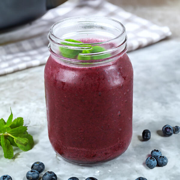 Ein Glas mit violettem Blaubeer-Eisshake steht auf einer Marmoroberfläche. Oben schwimmen zwei Minzblätter. Um das Glas liegen frische Blaubeeren und Minzblätter.
