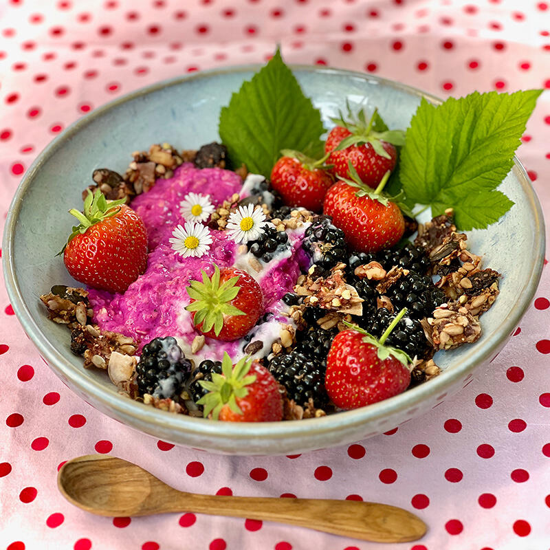 Gute-Laune-Beeren-Bowl mit homemade Granola Schale mit knall-pinkem veganem Joghurt, frischen Erdbeeren, Brombeeren, selbstgemachtem Granola und grünen Blättern. Drei kleine weiße Blüten liegen auf dem Joghurt. Unterlage ist eine rosa Tischdecke mit roten Punkten.
