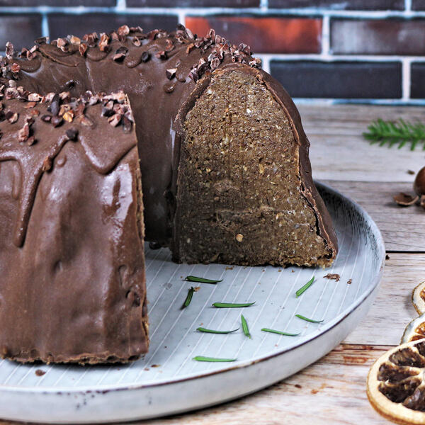 Weihnachtlicher Lebkuchen Gugelhupf Ein roh-veganer Lebkuchen Gugelhupf mit Schokoladenglasur und Nussstückchen wird auf einem Teller präsentiert. Ein Stück des Kuchens ist aufgeschnitten und zeigt die dichte, gewürzte Innenstruktur.