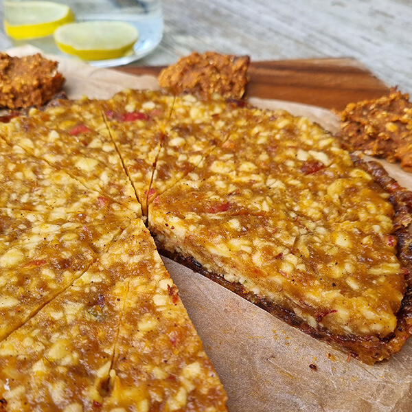 Rohe Apfel -Tarte Rohe Apfel-Tarte mit sichtbaren Apfelstücken und Zimt, in mehrere Stücke geschnitten. Sie liegt auf Backpapier auf einem Holzbrett. Im Hintergrund stehen Kekse und ein Glas Wasser mit Zitronenscheiben.