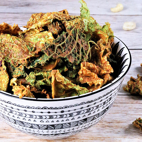 Smokey Limetten-Wirsingchips, roh-vegan Weiß-schwarz gemusterte Schale gefüllt mit getrockneten, grünen und braunen Wirsingchips auf hellem Holztisch. Einige Chips und Cashewnüsse liegen daneben verstreut.