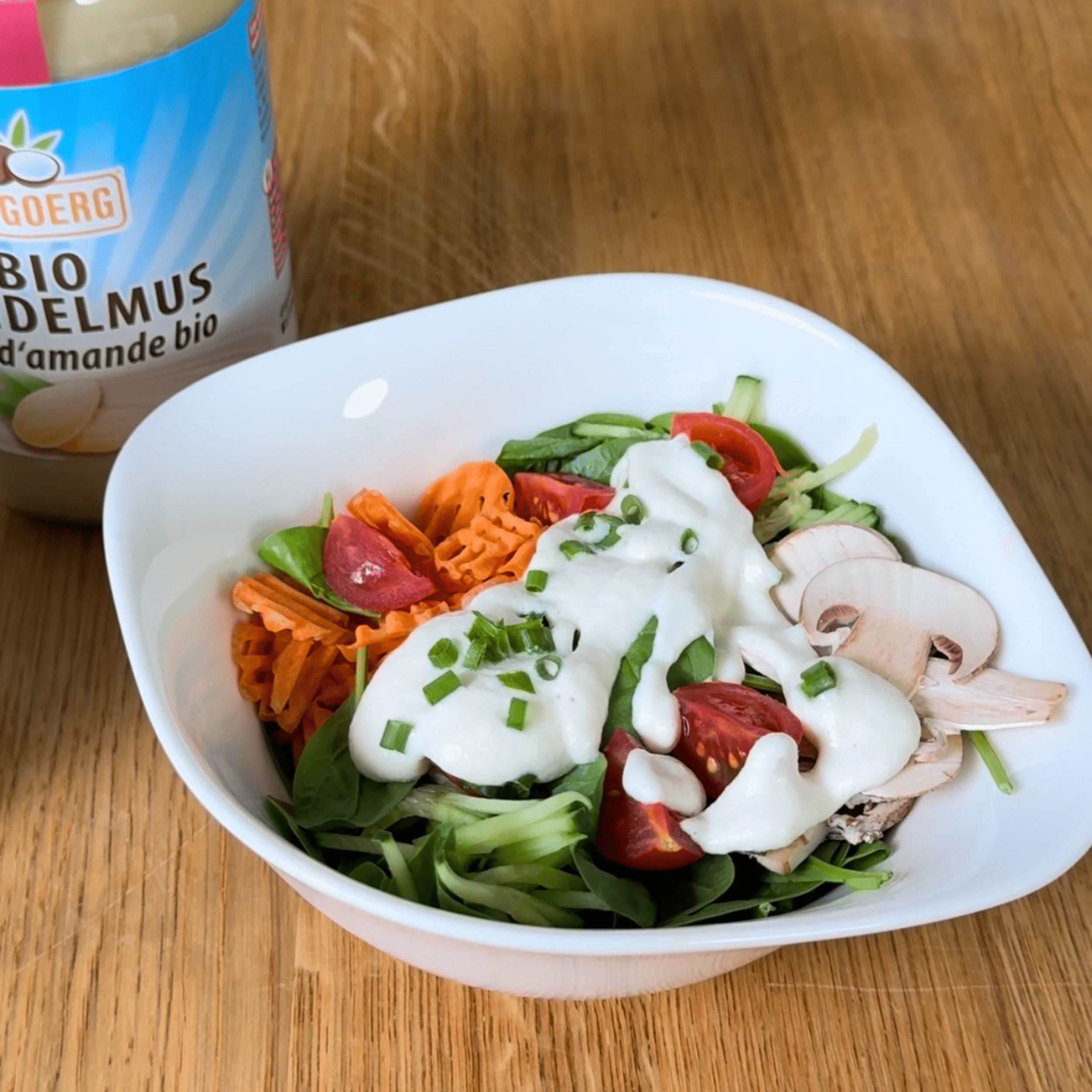 Schnittlauch Salatdressing Weiße Schüssel mit grünem Salat, Kirschtomaten, gewellten Karottenscheiben, rohen Champignons und weißem Schnittlauch-Salatdressing. Daneben steht ein Glas mit Bio-Mandelmus.