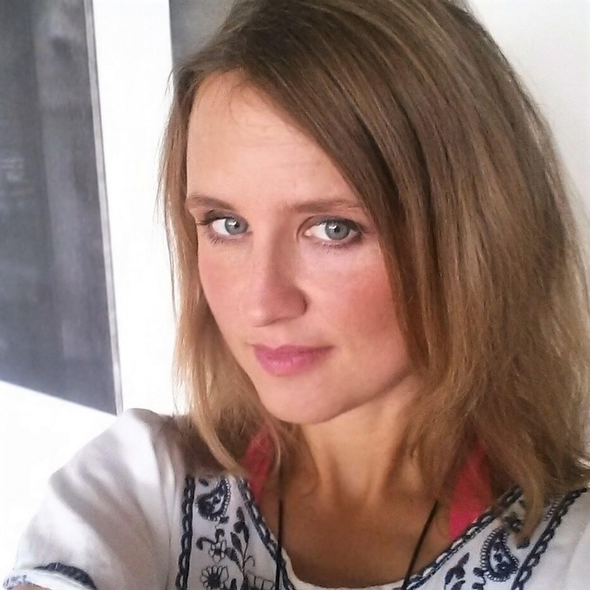 Angie schaut direkt in die Kamera. Sie hat schulterlanges, braunes Haar und trägt ein weißes Oberteil mit blauem Muster. Der Hintergrund ist unscharf.