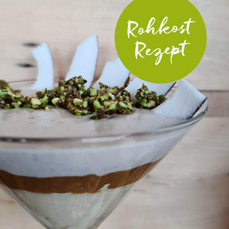Glas mit geschichteter Mango-Ananas-Kokos-Creme. Oben sind Kokosstückchen und grüne Saaten als Topping verteilt. Text Rohkost Rezept in grünem Kreis.