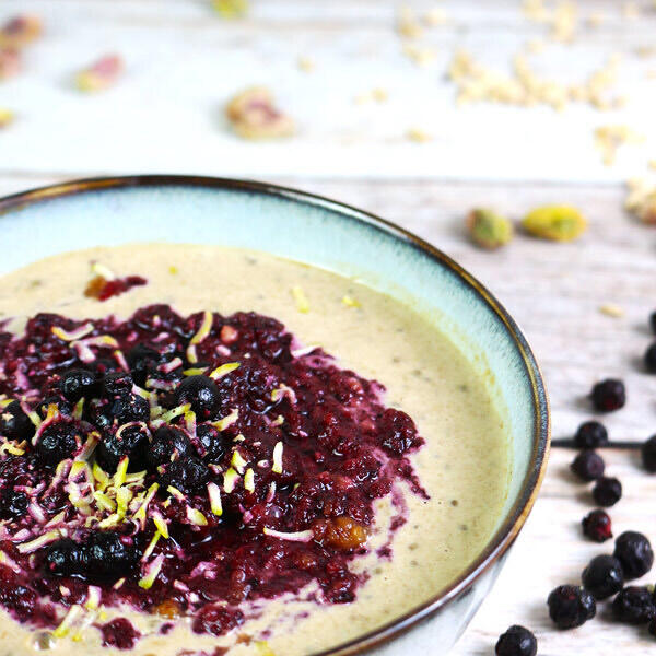 Blaubeer-Pistazien-Porridge, roh-vegan Schüssel mit cremigem rohem, veganem Porridge, garniert mit dunklem Waldblaubeerenmus und gehackten Pistazien. Im Hintergrund sind verstreute Pistazien und Blaubeeren auf Holzoberfläche zu sehen.