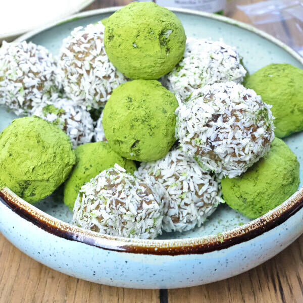 Matcha Energieballs Eine Schale mit runden Energieballs, einige bedeckt mit grünem Matcha-Pulver, andere mit Kokosraspeln, alle eng aneinandergelegt auf einem Holztisch.