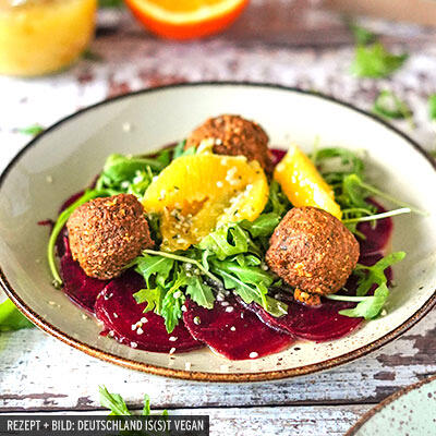 Drei braune Gemüse-Nussbällchen liegen auf einem Teller mit dünnen roten Rote-Bete-Scheiben, grünen Rucolablättern und gelben Orangenscheiben.