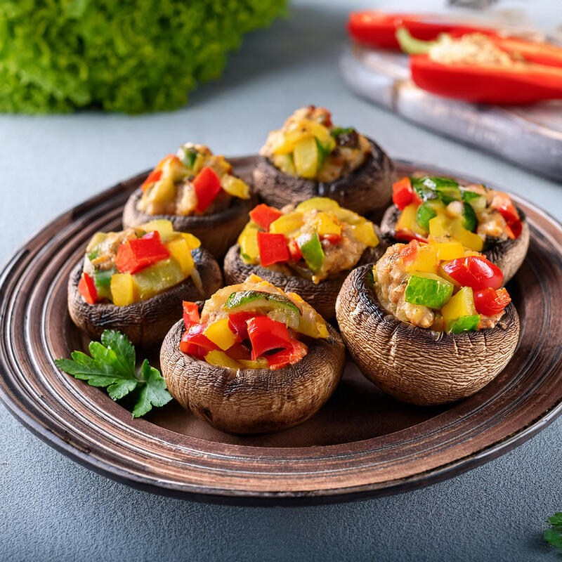Gefüllte Steinchampignons, roh-vegan von Peter Dreverhoff Sieben gefüllte Steinchampignons mit einer bunten Mischung aus roten, gelben und grünen Paprikastücken liegen auf einem braunen Teller.