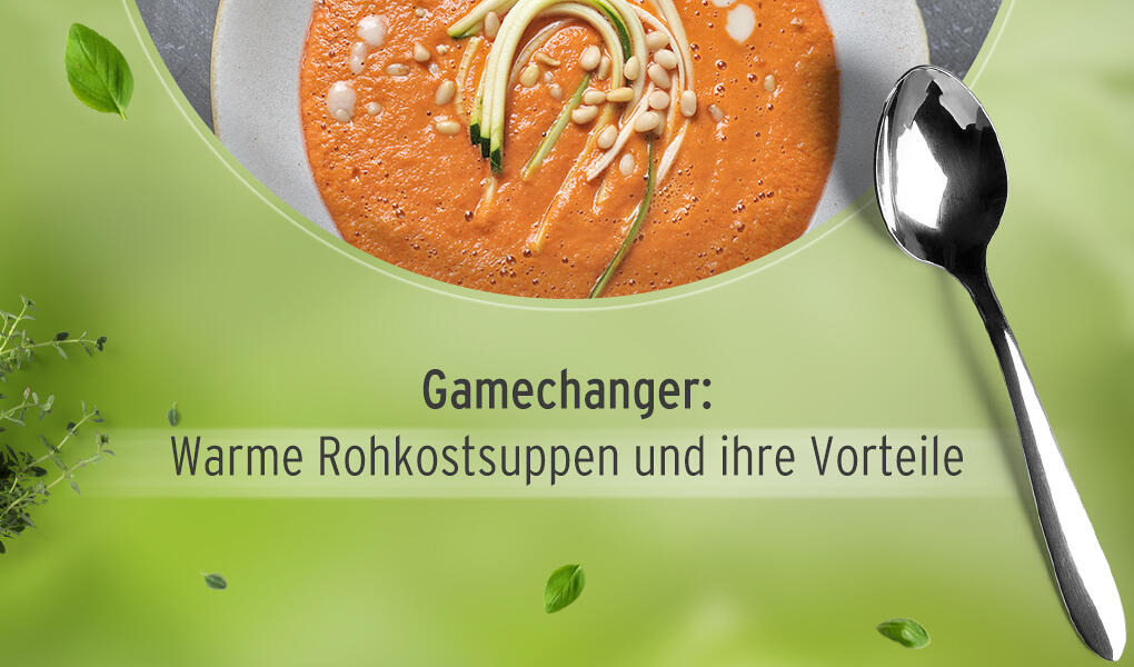 Orangefarbene Suppe mit Zucchinistreifen, Pinienkernen und kleinen weißen Tropfen auf einem weißen Teller. Rechts daneben liegt ein glänzender silberner Löffel auf grünem Hintergrund.