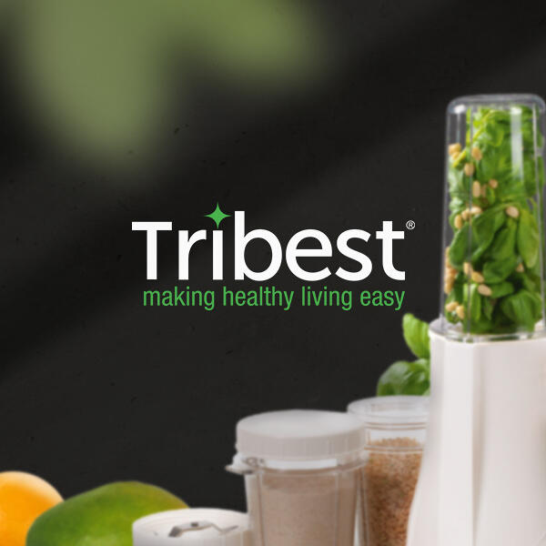 Der Tribest-Mixer ist mit frischem Spinat und Nüssen befüllt. Im Vordergrund stehen verschiedene Zutaten und Aufsätze vor einem dunklen Hintergrund. Das Tribest-Logo und der Slogan making healthy living easy sind zentral platziert.
