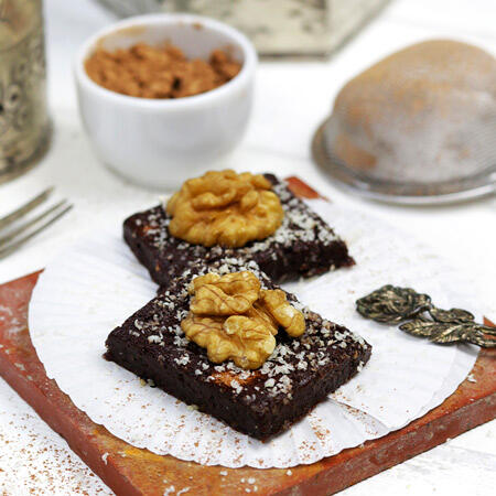 Zwei quadratische Schokoladenstücke liegen auf einem weißen Papierförmchen. Jedes Stück ist mit Walnusshälften und feinen weißen Streuseln garniert. Im Hintergrund steht eine kleine weiße Schüssel mit braunen Zutaten.