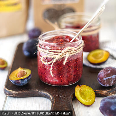 Ein Glas Zwetschgen-Chutney auf einem Holzbrett mit einer weißen Schnur umwickelt und einem Löffel darin. Rundherum liegen halbierte und ganze Zwetschgen.