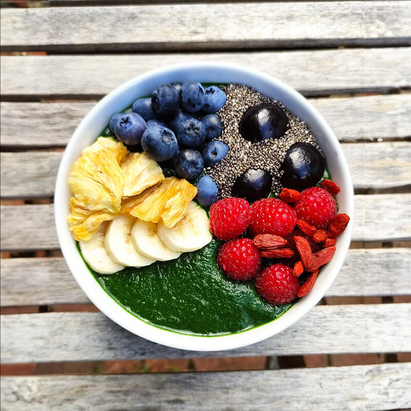 Keimling Olympic Smoothiebowl Schale mit grüner Smoothiebasis, belegt mit Ananasstücken, Bananenscheiben, Blaubeeren, Chiasamen, schwarzen Kirschen, Himbeeren und Gojibeeren auf einem Holztisch.