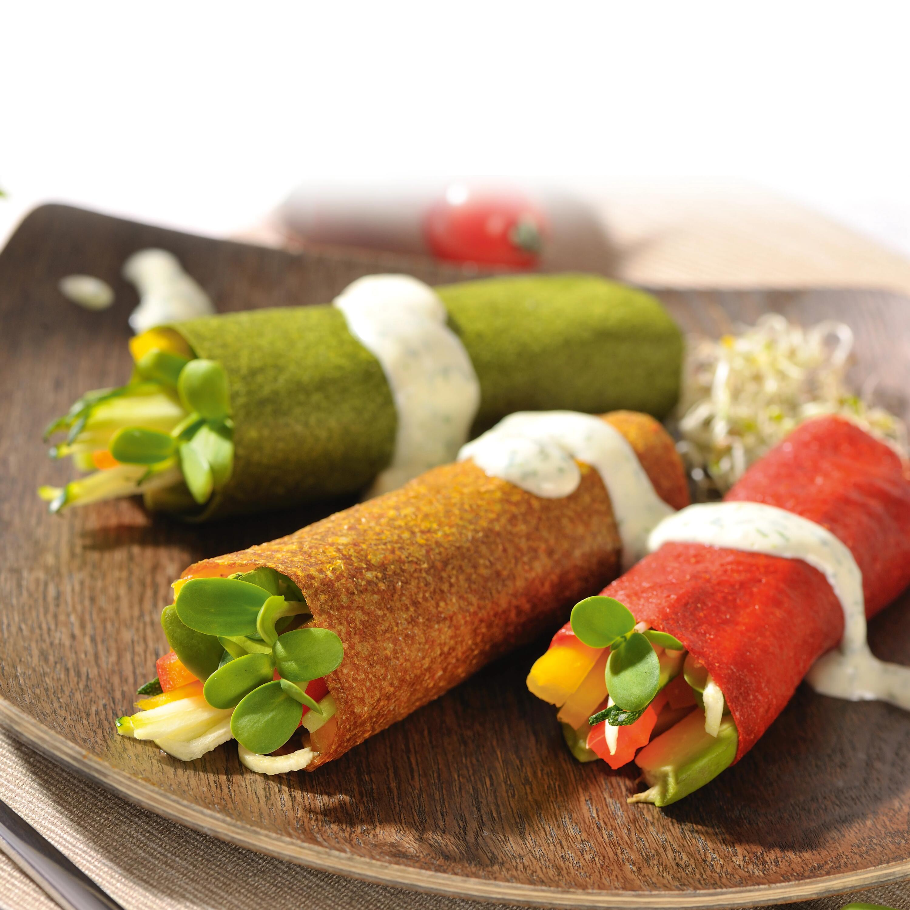Bunte Rohkost Wraps Drei farbige Gemüse-Wraps liegen auf einem Holzbrett. Jeder Wrap ist mit frischem Gemüse gefüllt und mit weißer Sauce garniert. Ein roter Cherrytomate liegt im Hintergrund.