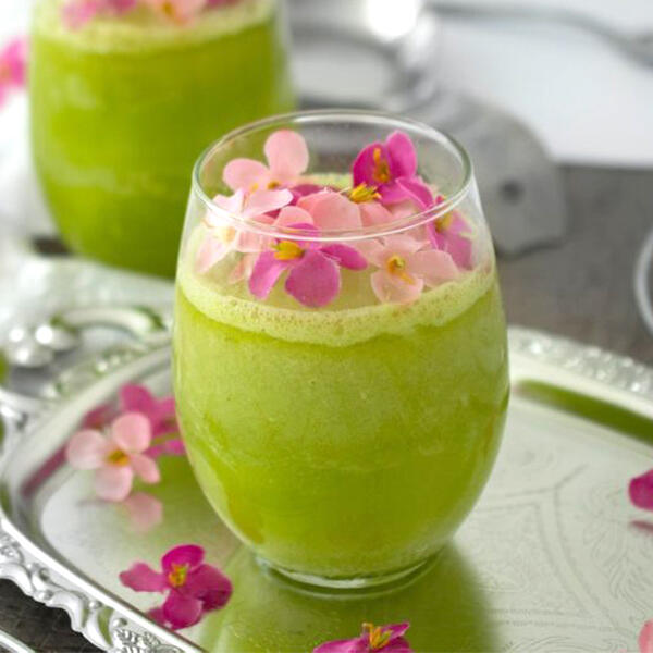 Endivien-Smoothie von Peter Dreverhoff Glas mit grünem Endivien-Smoothie steht auf einem silbernen Tablett. Der Smoothie ist mit rosa und pinken essbaren Blüten dekoriert. Im Hintergrund steht ein zweites Glas mit dem selben Getränk.