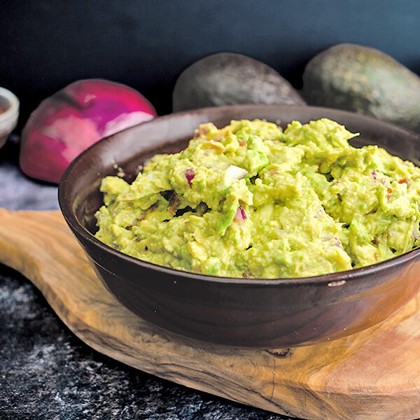 Die beste Guacamole - mit Kern! roh-vegan nach Peter Dreverhoff Schwarze Schale mit cremiger, stückiger Guacamole, erkennbar mit grünen Avocadostücken und roten Zwiebelstücken, steht auf einem Holzbrett vor einer roten Zwiebel und zwei Avocados.