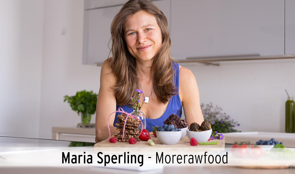 Maria Sperling sitzt lächelnd an einem Tisch in einer modernen Küche. Vor ihr liegen verschiedene kleine Schalen mit Beeren, Nüssen und Rohkost-Snacks.