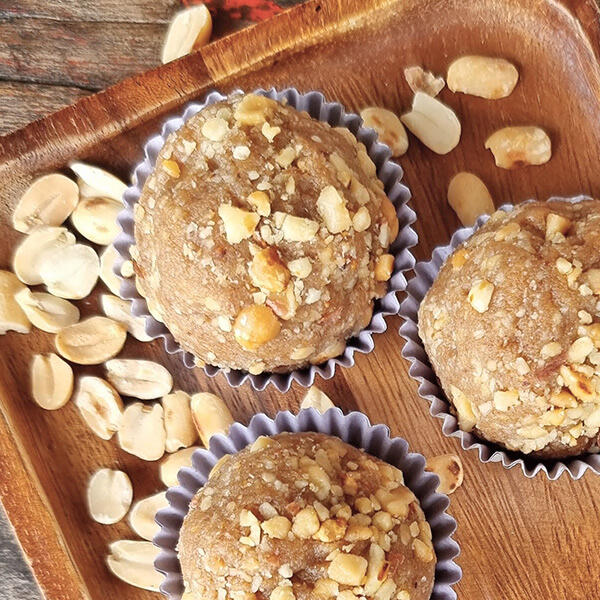 Protein Snacks Drei proteinreiche Snackbällchen liegen auf einem Holzbrett und sind mit gehackten Nüssen bestreut. Um sie herum liegen zusätzliche Erdnussstücke.
