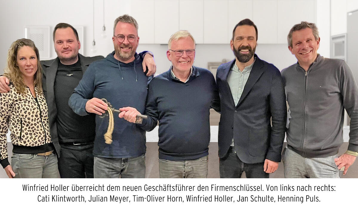 Winfried Holler überreicht Tim-Oliver Horn symbolisch den Firmenschlüssel. Sechs Personen stehen in einer Reihe und lächeln in die Kamera.