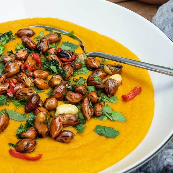 Karotten-Kokos-Linsensuppe mit feurigen Koriander-Erdnüssen Eine Schale mit cremiger oranger Karotten-Kokos-Linsensuppe, garniert mit gerösteten Erdnüssen, frischem Koriander und roten Chilistreifen, daneben liegt ein silberner Löffel.