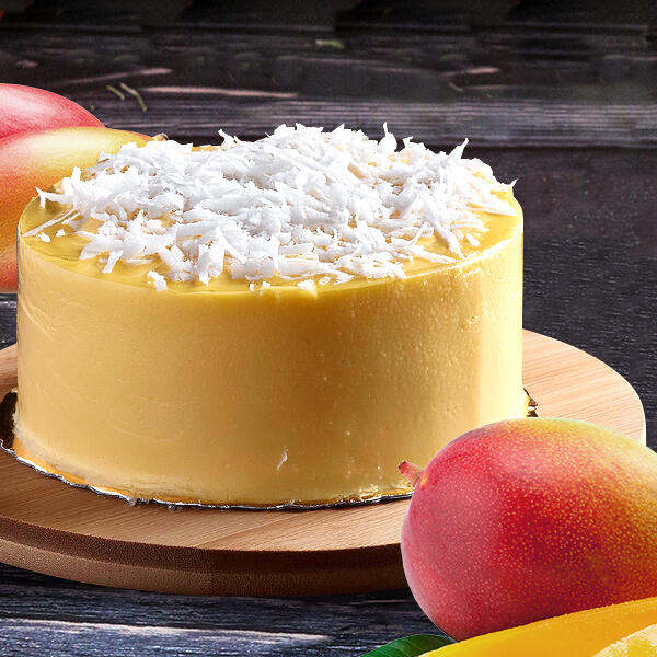 Raw Mango Cake von Peter Dreverhoff Runder, gelber Roh-veganer Mangokuchen mit weißer Kokosraspel-Dekoration auf einem Holzbrett. Zwei ganze Mangos liegen daneben auf einer dunklen Oberfläche.