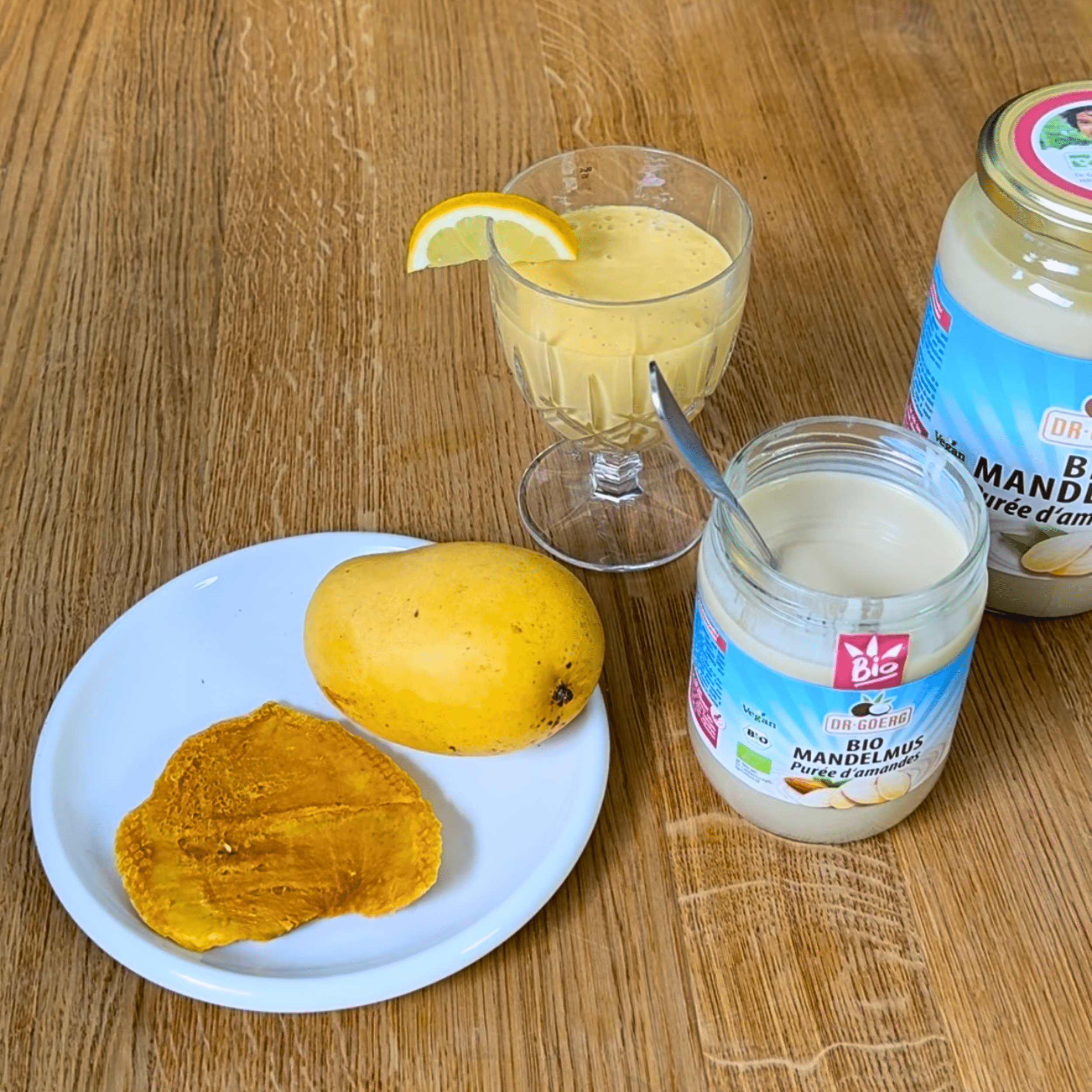 Mango Mandel Lassi Ein Glas Mango Mandel Lassi mit Zitronenscheibe am Rand steht neben einer ganzen Mango und getrockneten Mangoscheiben auf einem Teller. Daneben steht ein geöffnetes Glas Bio Mandelmus mit einem Löffel.