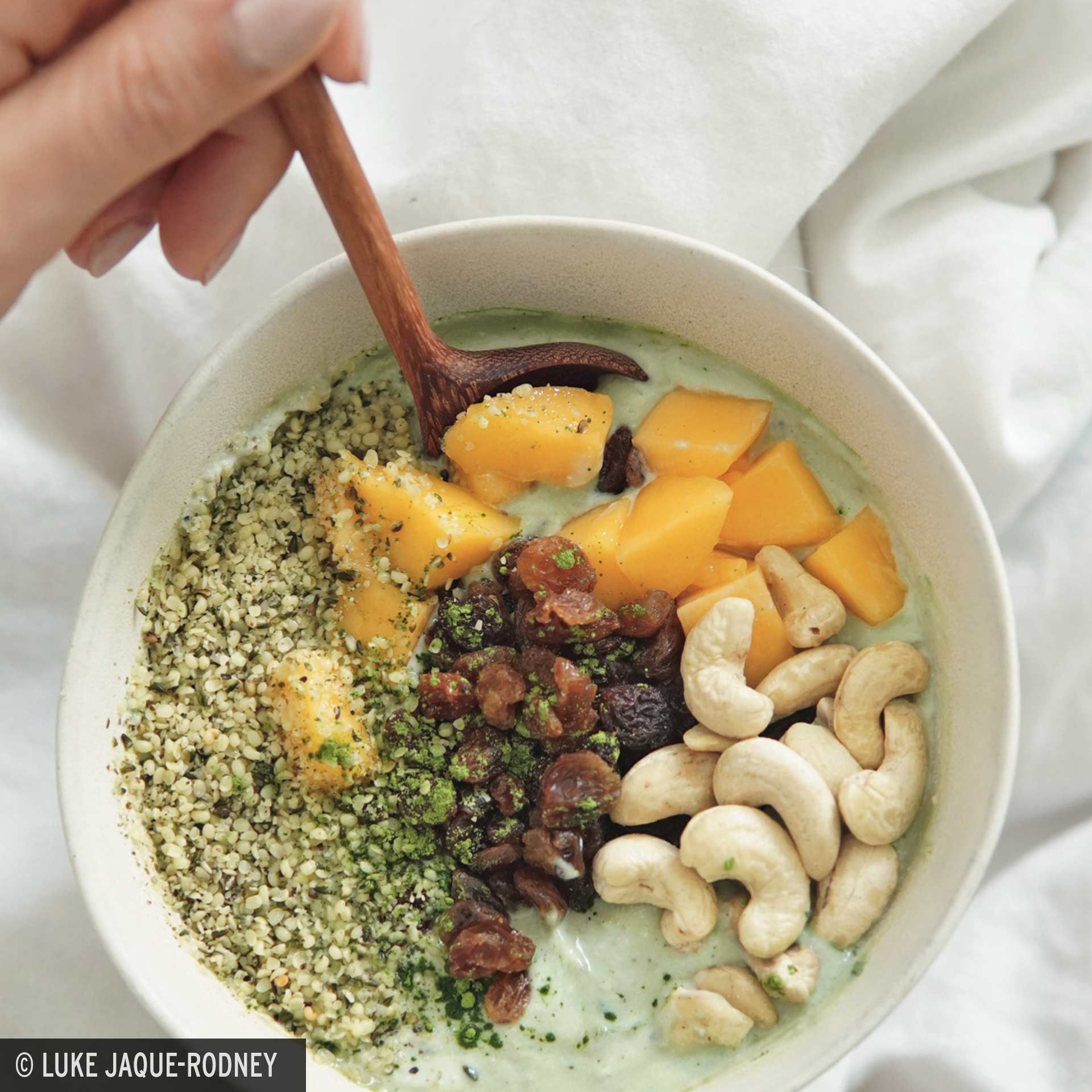 Luke´s Rezept für Matcha Dream Eine Schüssel mit pflanzlichem Joghurt, Banane, Mango, Hanfsamen, Cashewkernen, Rosinen und grünem Matcha-Pulver. Eine Hand hält einen Holzlöffel in der Schüssel.