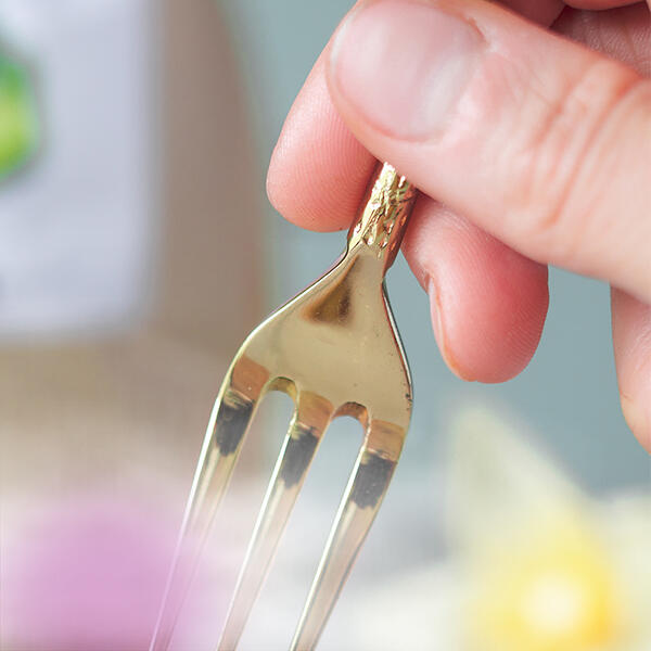 Eine Hand hält eine goldfarbene Gabel mit drei Zinken vor einem unscharfen, pastellfarbenen Hintergrund.