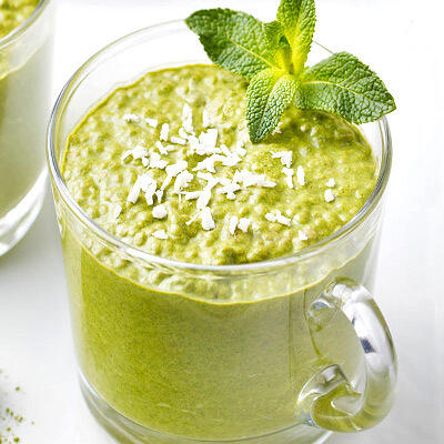 Chiapudding mit Matcha Glas mit grünem Chiapudding, garniert mit Kokosraspeln und frischem Minzblatt auf weißem Untergrund.