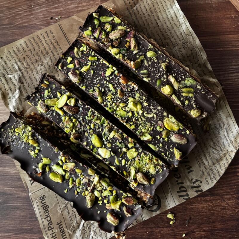 Pistachio Bars Vier rechteckige Pistazienriegel mit dunkler Schokolade und grob gehackten Pistazienstücken liegen auf einem Stück Zeitungspapier auf einem Holztisch.
