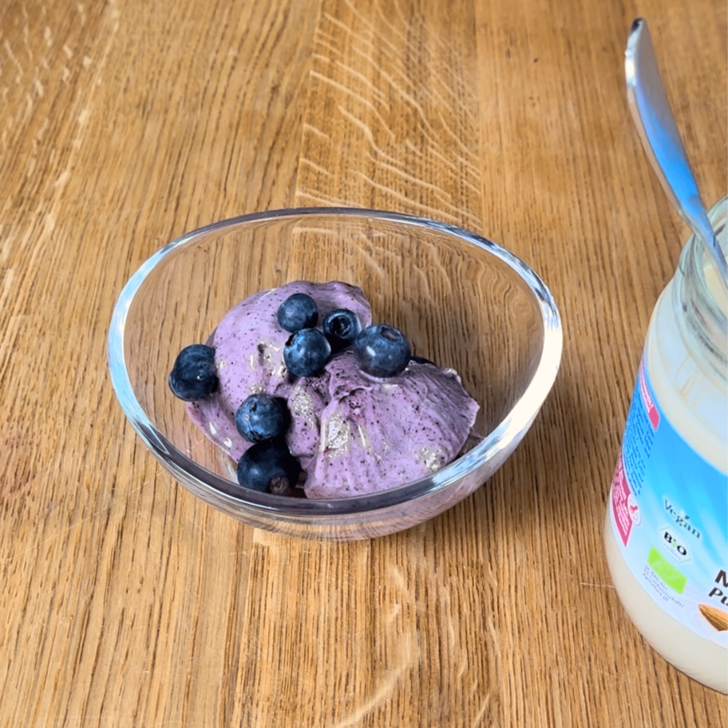 Mandel Beeren Eiscreme Zwei Kugeln lila Mandel-Beeren-Eiscreme mit frischen Wildheidelbeeren in einer Glasschale auf einem Holztisch. Daneben steht ein Glas Mandelmus mit einem Metalllöffel.