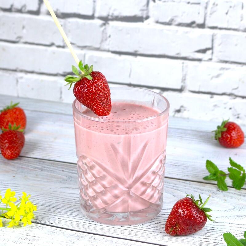 Erdbeer Lassi, roh-vegan von Angela Griem Ein Glas mit rosa Erdbeer-Lassi steht auf einem weißen Holztisch. Eine frische Erdbeere steckt am Rand des Glases auf einem Holzstab. Um das Glas liegen mehrere Erdbeeren und einige grüne Minzblätter.